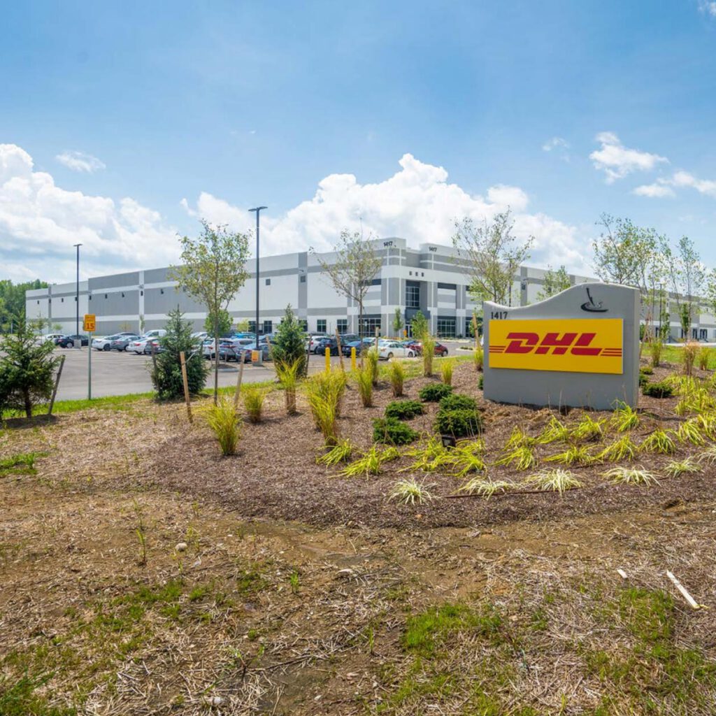 A large DHL warehouse and distribution center with a prominent DHL sign in front, surrounded by landscaped greenery and a parking lot filled with vehicles under a blue sky.