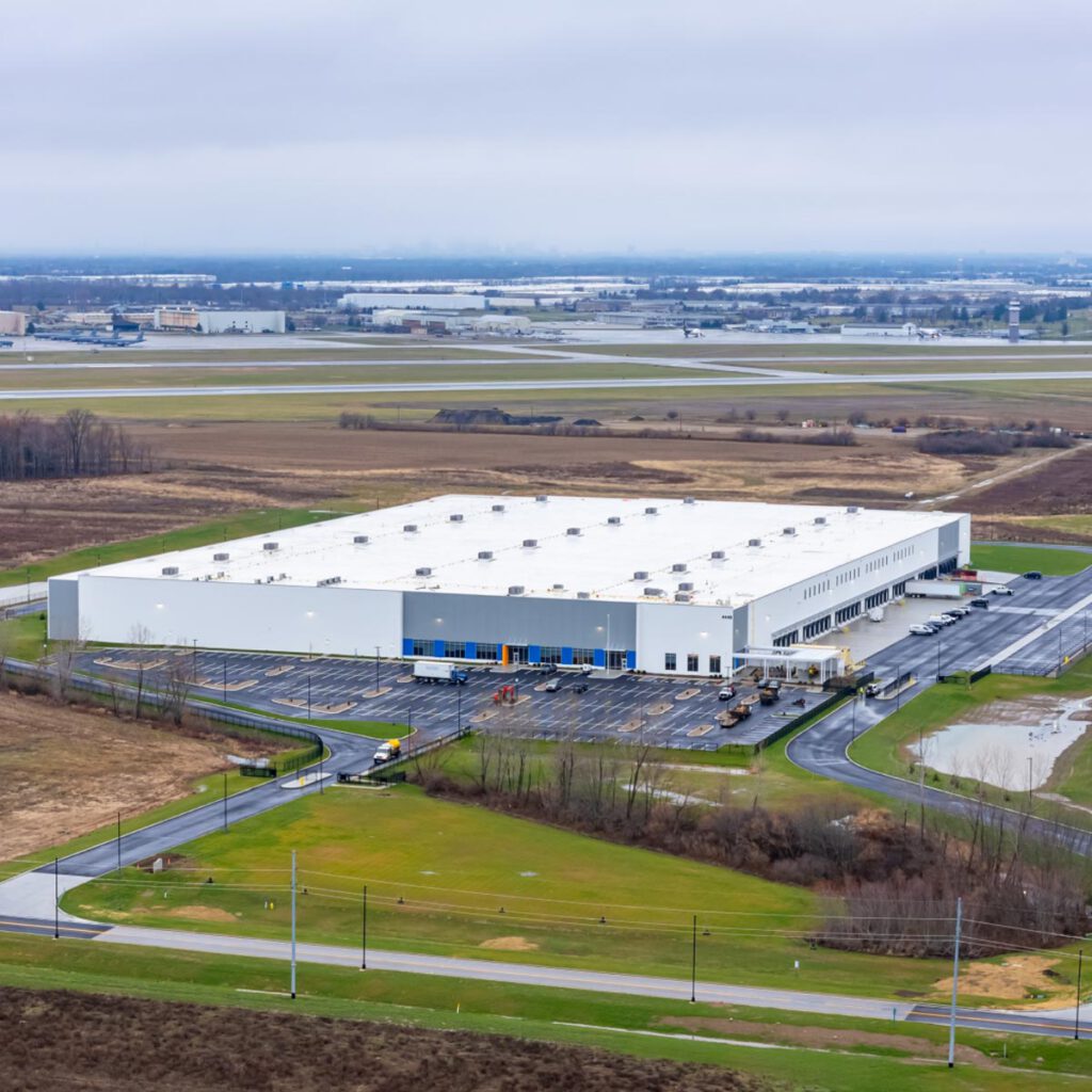Aerial view of a large, white industrial warehouse surrounded by parking lots, trucks, and green fields, with an airport runway and additional buildings visible in the background under a cloudy sky.