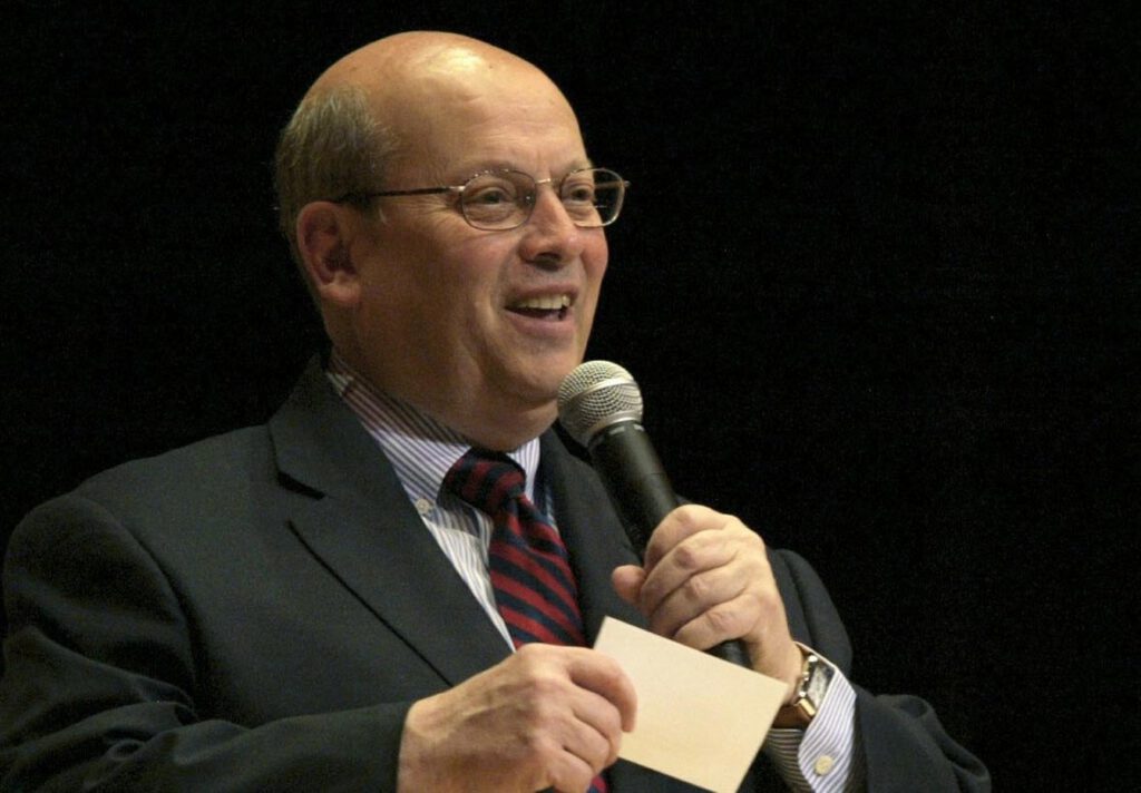 A man in a suit and striped tie smiles while speaking into a microphone. He holds a small white card in his other hand. The background is dark, highlighting the speaker.