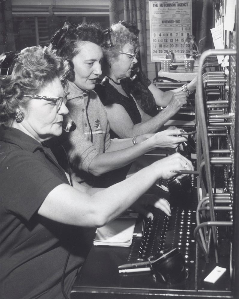 Three women sit side by side at a vintage telephone switchboard, wearing headsets and connecting calls by inserting cables. A calendar on the wall indicates the month of November.
