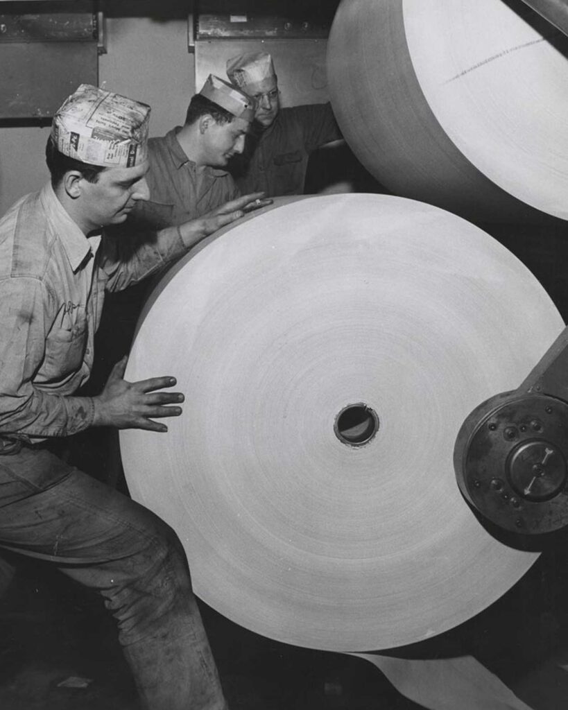 Three men wearing paper hats and work uniforms inspect a large roll of paper on an industrial machine in a factory setting. The men appear focused on the machinery and the paper roll.