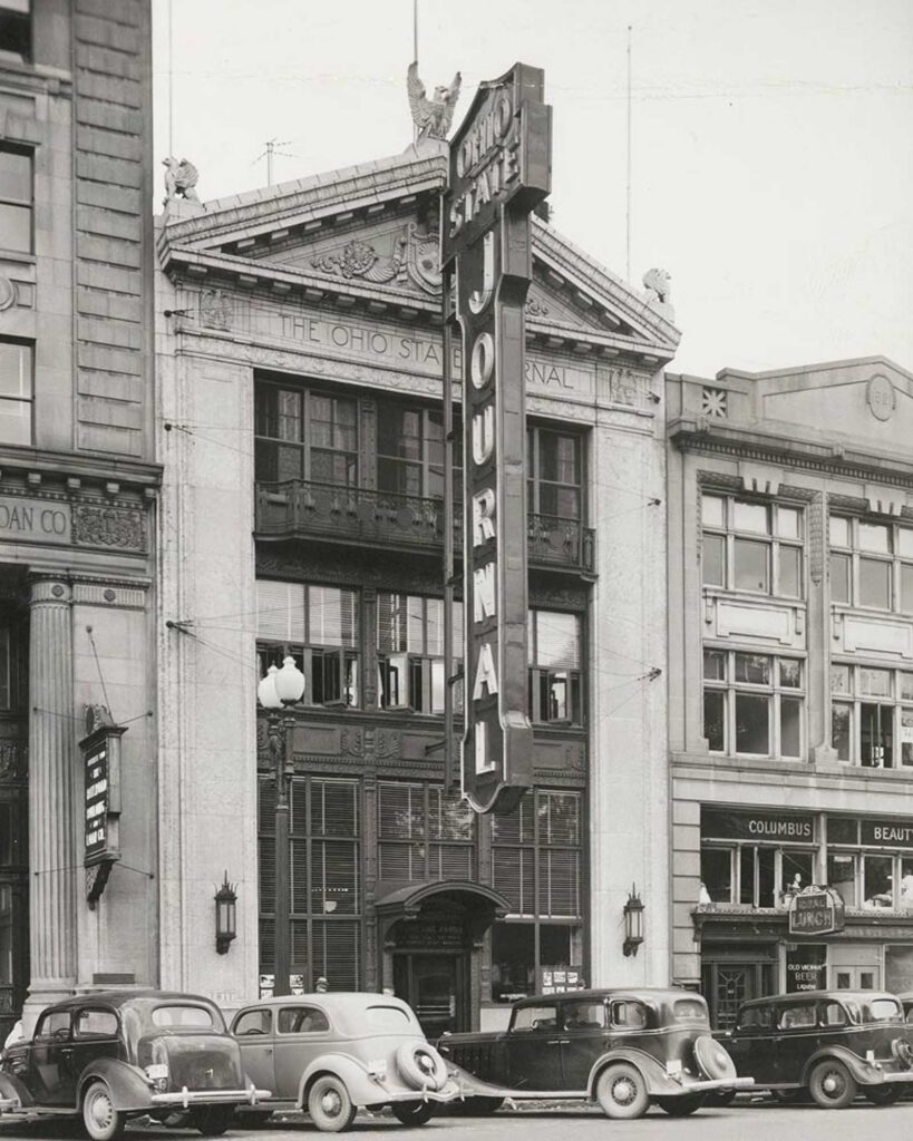 Black-and-white photo of The Ohio State Journal building, featuring a large vertical sign reading OHIO STATE JOURNAL. Several vintage cars are parked along the street in front of the historic office facade.