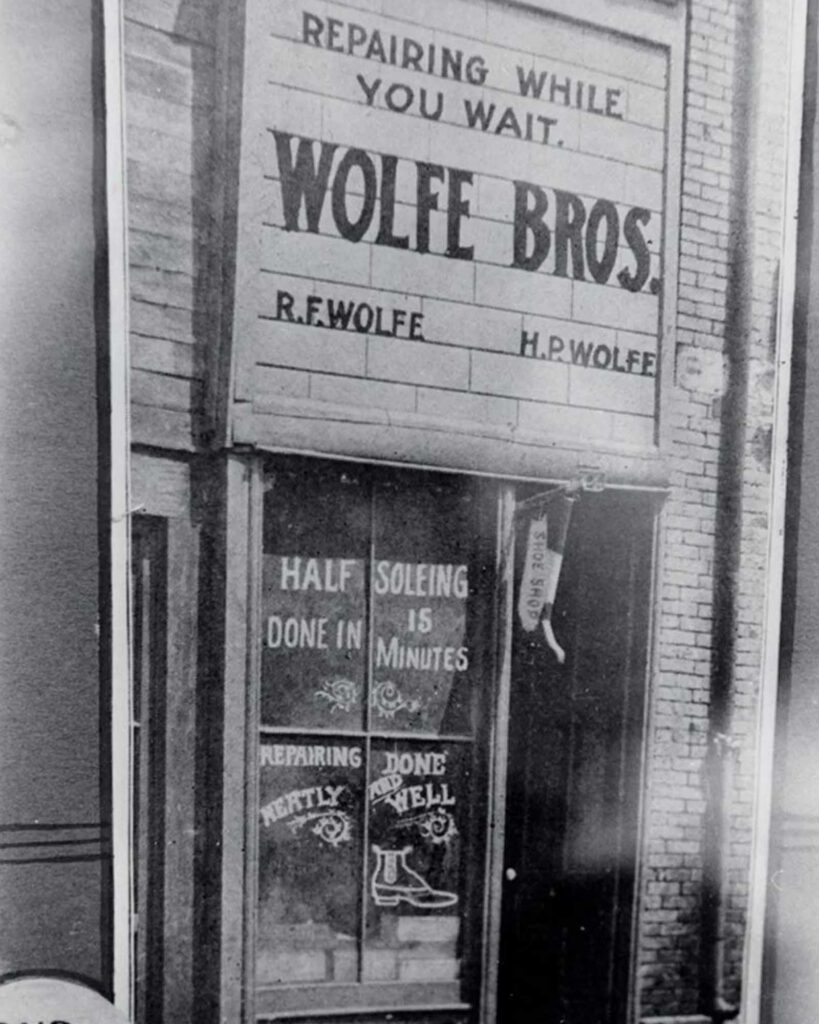 Black-and-white photo of a storefront with signs reading Wolfe Bros. and Repairing While You Wait. The window advertises shoe repairing and resoling, with illustrations of shoes and boots.