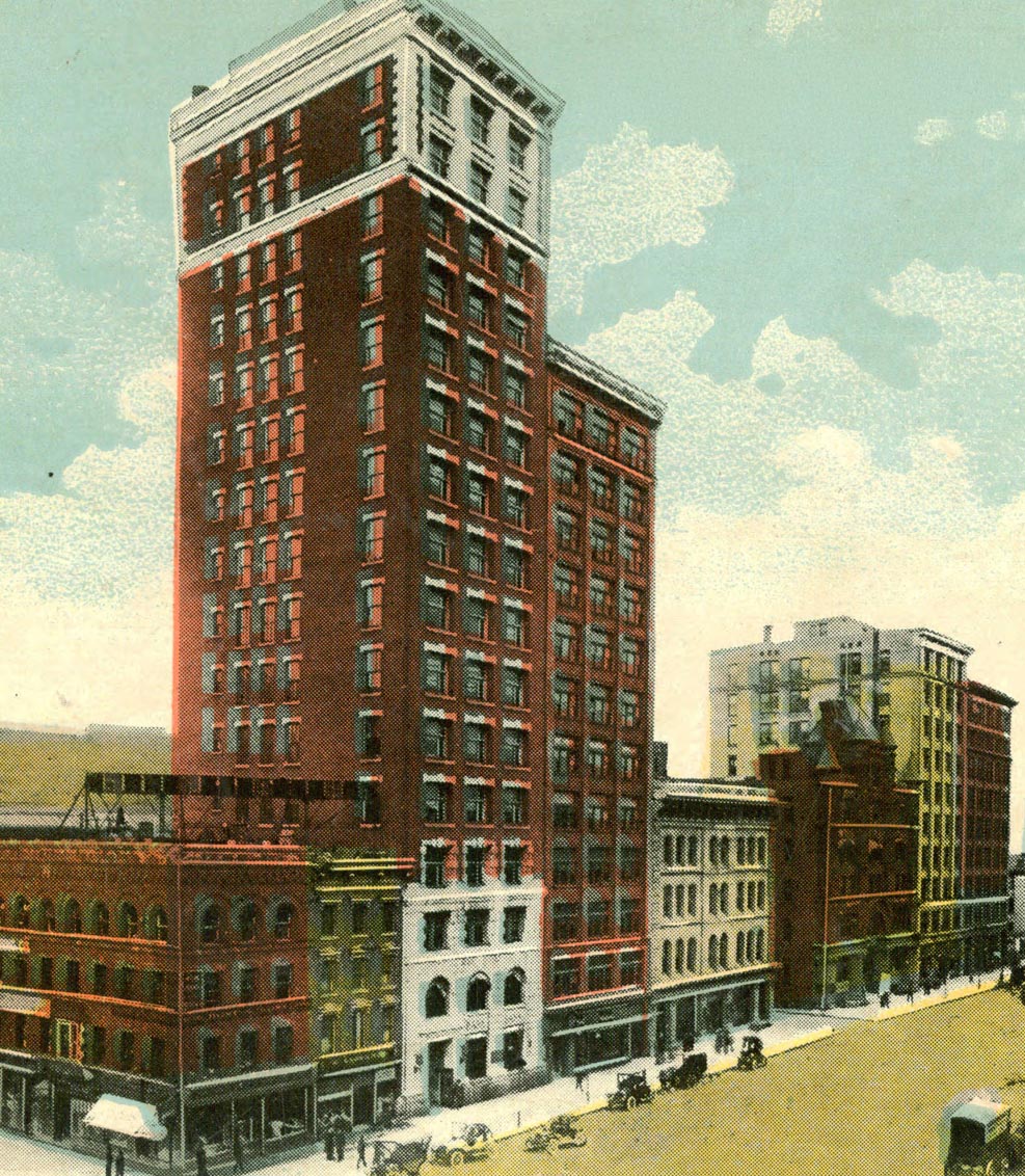 A tall red-brick skyscraper with white accents stands among shorter buildings on a busy city street with early 20th-century cars and people under a blue sky with scattered clouds.