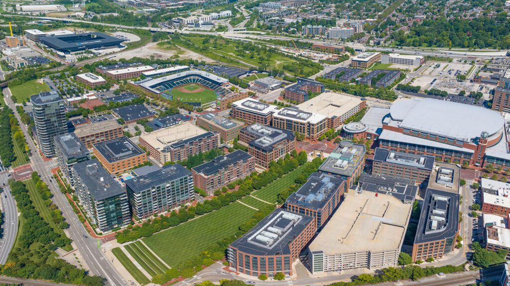 Aerial view of a large urban campus with multiple brick buildings, a green lawn, a stadium, and parking structures, surrounded by roads, trees, and residential areas in the background.