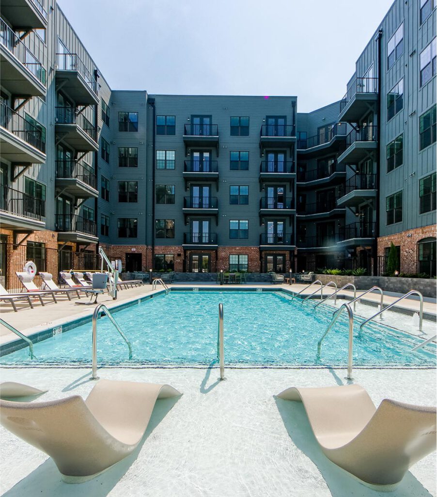 Apartment complex courtyard with a swimming pool, lounge chairs in shallow water, and multiple stories of balconies overlooking the pool area. Sunny, modern, and inviting atmosphere.