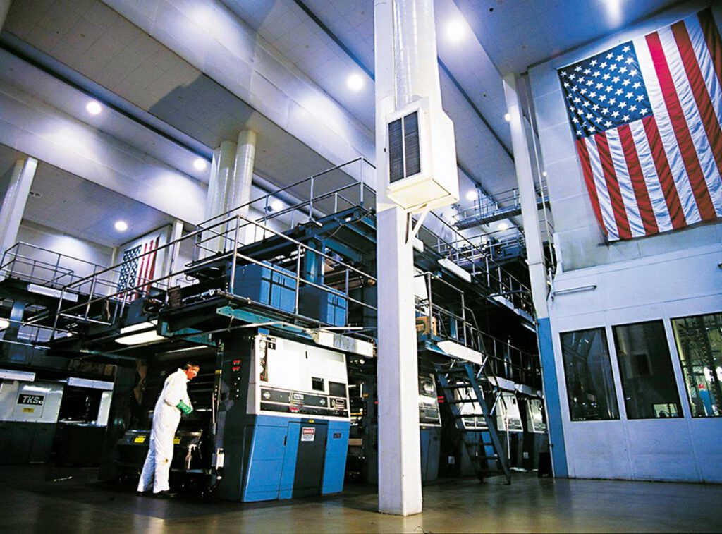 A worker in white overalls operates large industrial printing machines inside a spacious, well-lit facility. An American flag hangs prominently on the right wall above a glass-enclosed office area.