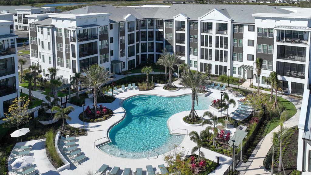 Aerial view of a modern apartment complex with a large, curvy outdoor swimming pool surrounded by lounge chairs, palm trees, and landscaped gardens. Four-story buildings encircle the pool area.