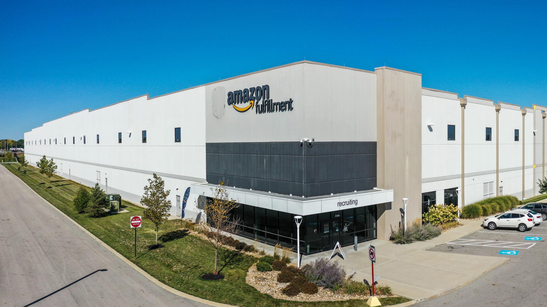 A large Amazon Fulfillment Center with a white exterior, company logo, and parking lot, seen on a clear, sunny day with several cars parked and some greenery around the building.