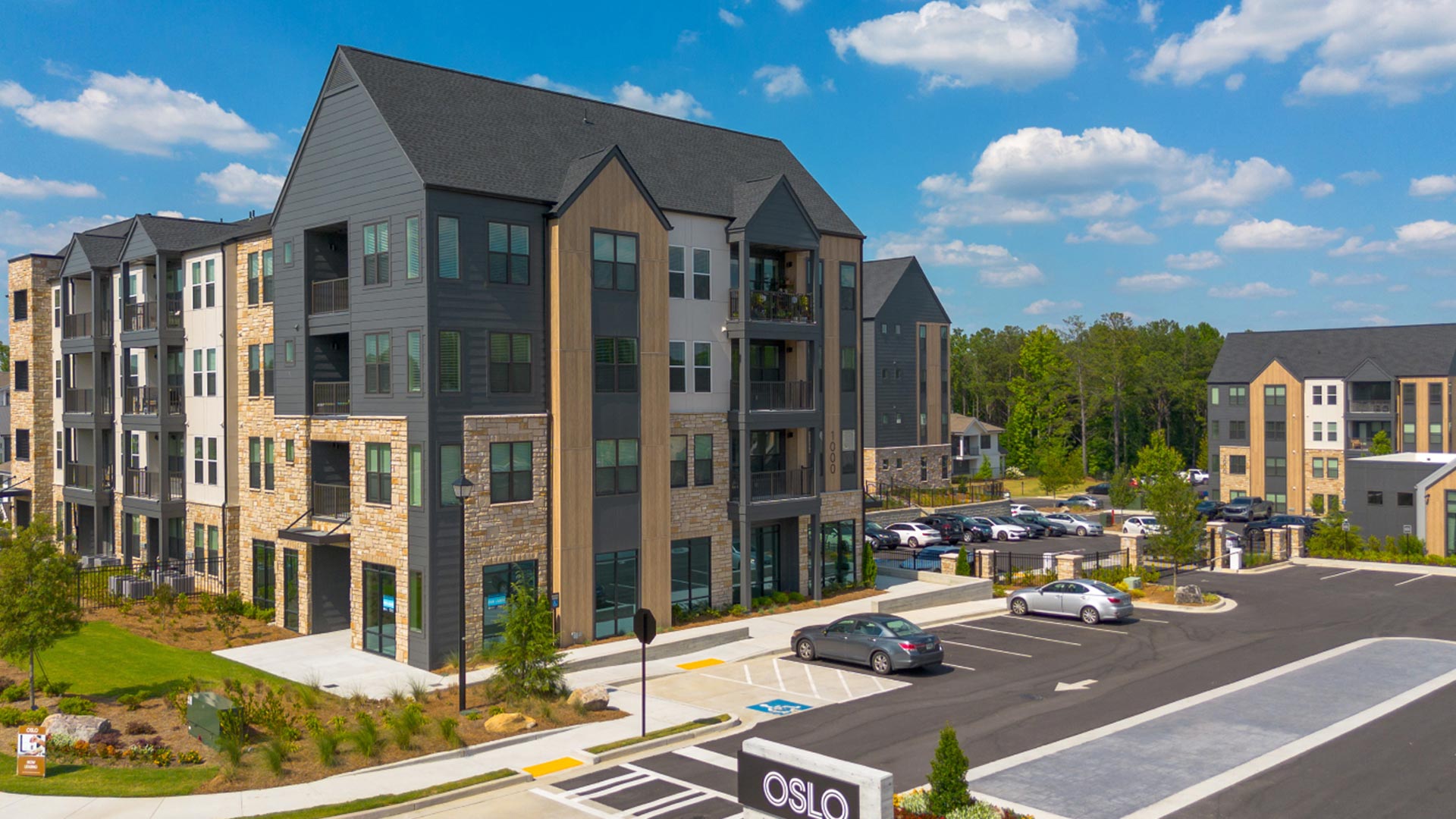 A modern apartment complex with dark and light exterior panels, large windows, and stone accents, surrounded by parked cars, trees, and a clear blue sky. A sign with OSLO is visible in the parking lot.