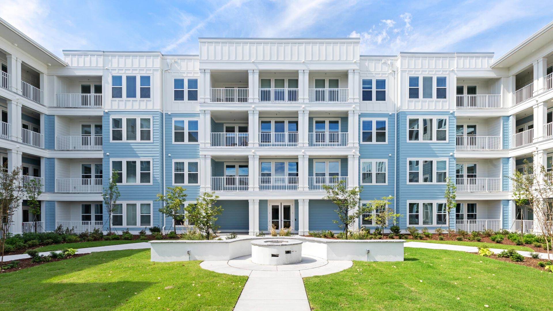 Four-story modern apartment building with light blue siding and white trim, featuring balconies, large windows, and a well-manicured lawn with a circular stone planter and a walkway in the foreground under a blue sky.