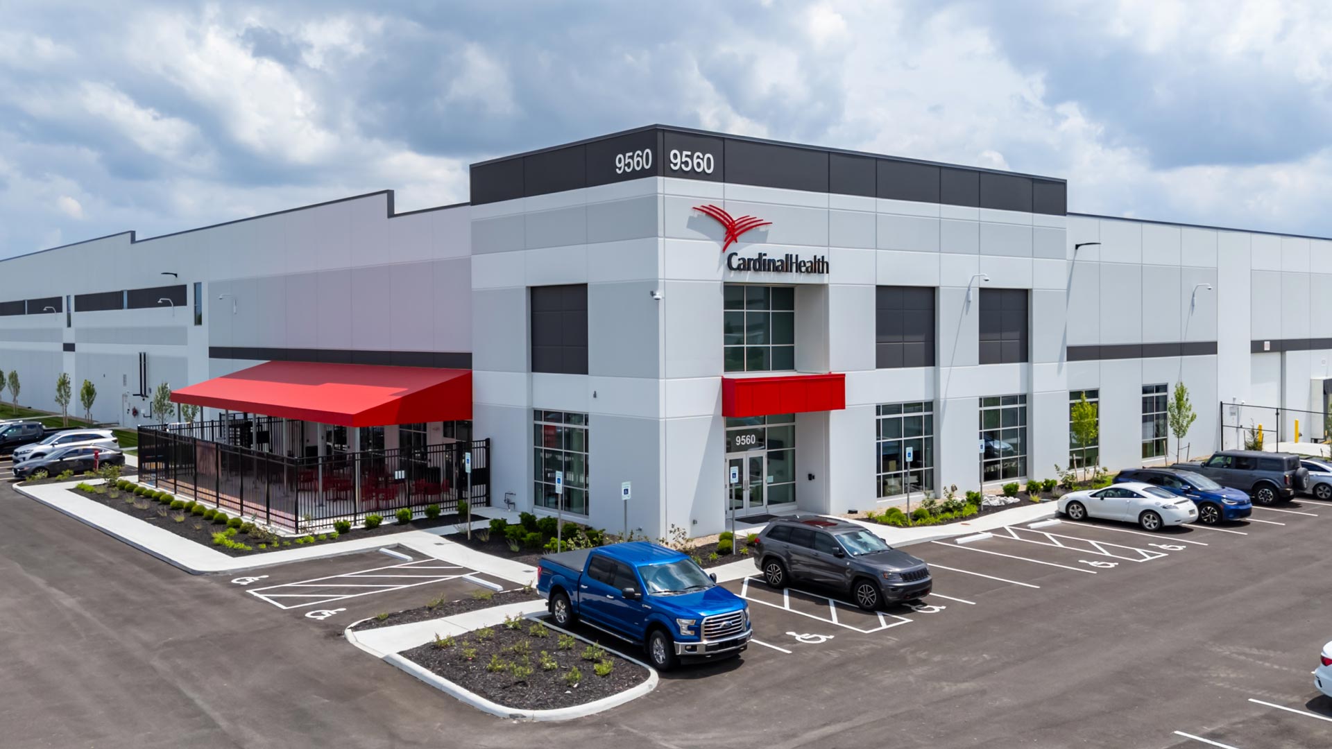 A modern warehouse building with Cardinal Health signage, large windows, and a red awning over an outdoor seating area. Several cars are parked in the lot, and the sky is partly cloudy.