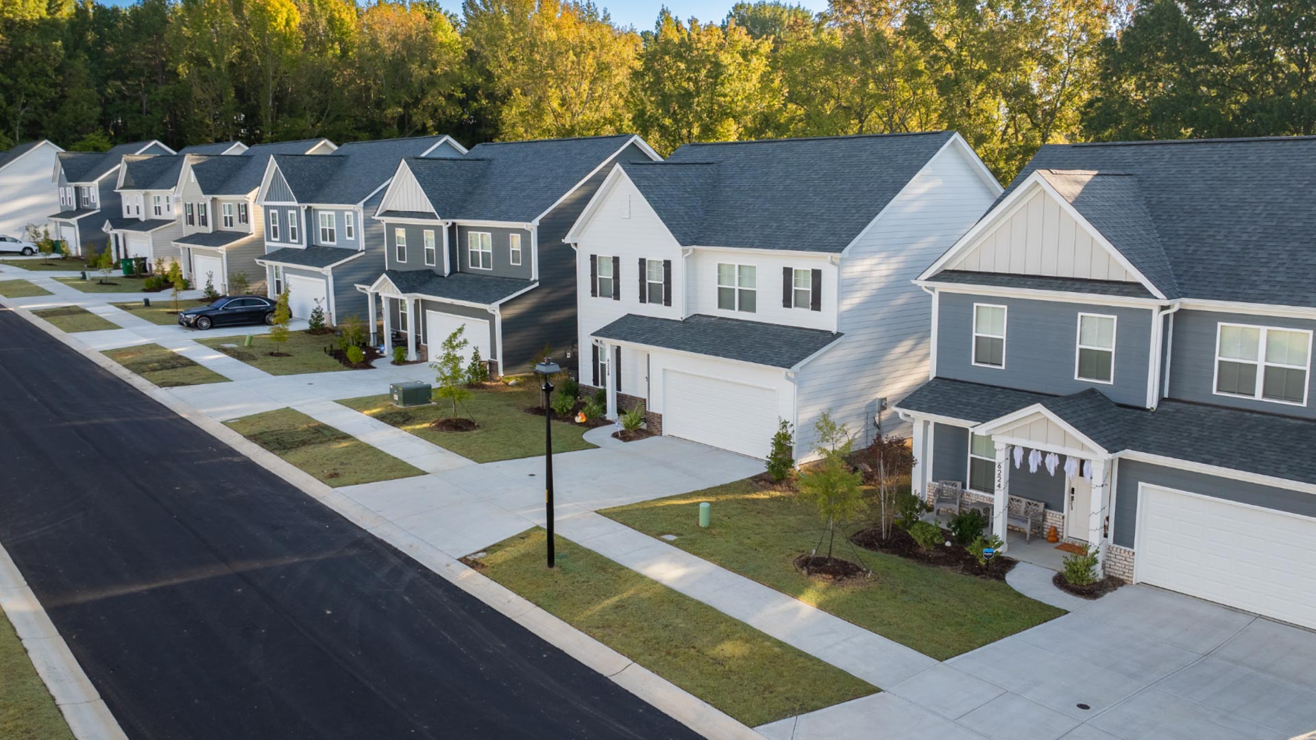 A row of modern two-story suburban houses with garages lines a clean, quiet street. Each home has a small lawn and driveway, with trees and greenery in the background under clear, sunny skies.