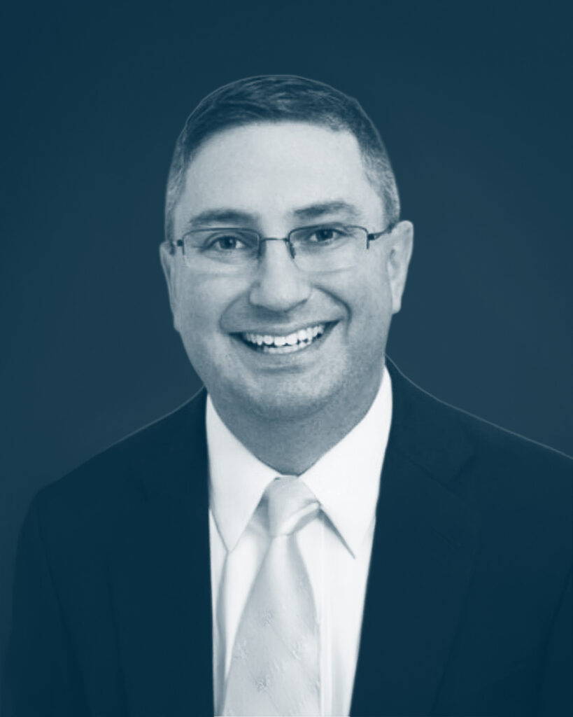 A man wearing glasses, a suit jacket, a white dress shirt, and a light-colored tie smiles at the camera against a plain, dark background.