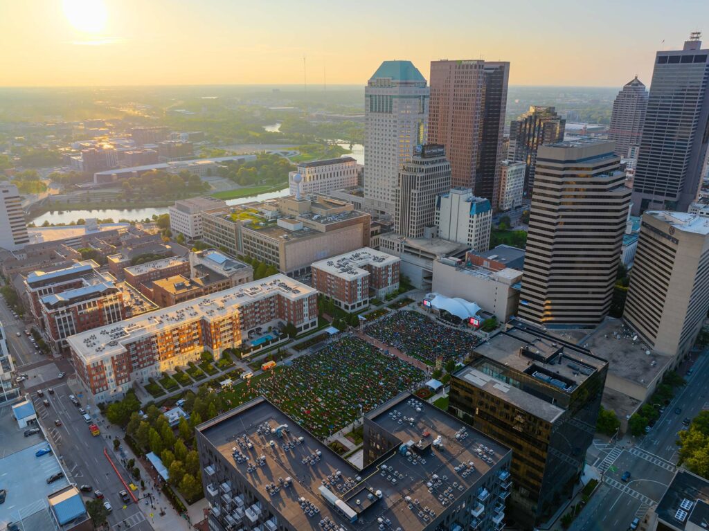 Aerial view of a city at sunset, with tall buildings, a river in the background, and a large outdoor gathering or event in a plaza surrounded by skyscrapers. The sunlight casts a warm glow over the scene.