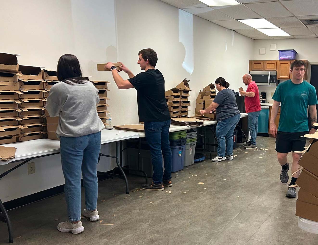 Five people are assembling cardboard boxes at tables in a room. Some are stacking finished boxes, while others fold them. The floor is strewn with cardboard scraps, and a kitchen area is visible in the background.