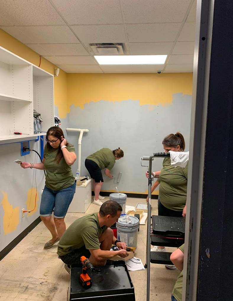 Four people wearing green shirts are painting and working in a small, partially renovated room with yellow and gray walls. Supplies and tools are scattered around as they work together on the project.