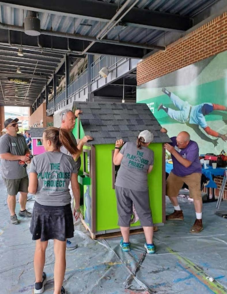 Five people work together to assemble a small, brightly colored playhouse under a covered outdoor area. Some wear The Playhouse Project shirts and tools are visible nearby. A mural is on the brick wall behind them.