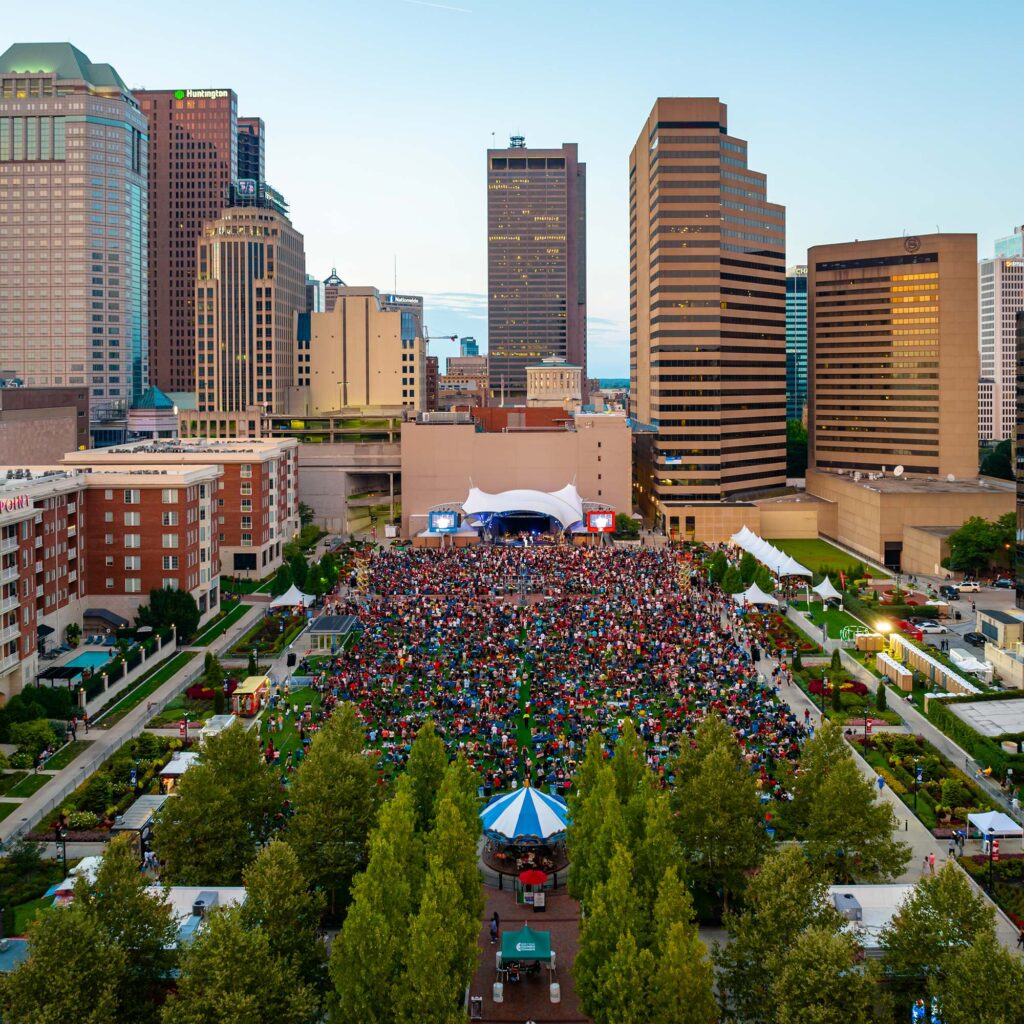 An aerial view of a large outdoor concert in a city park, with a crowd gathered on the lawn facing a stage. Tall buildings surround the park, and trees line the foreground.