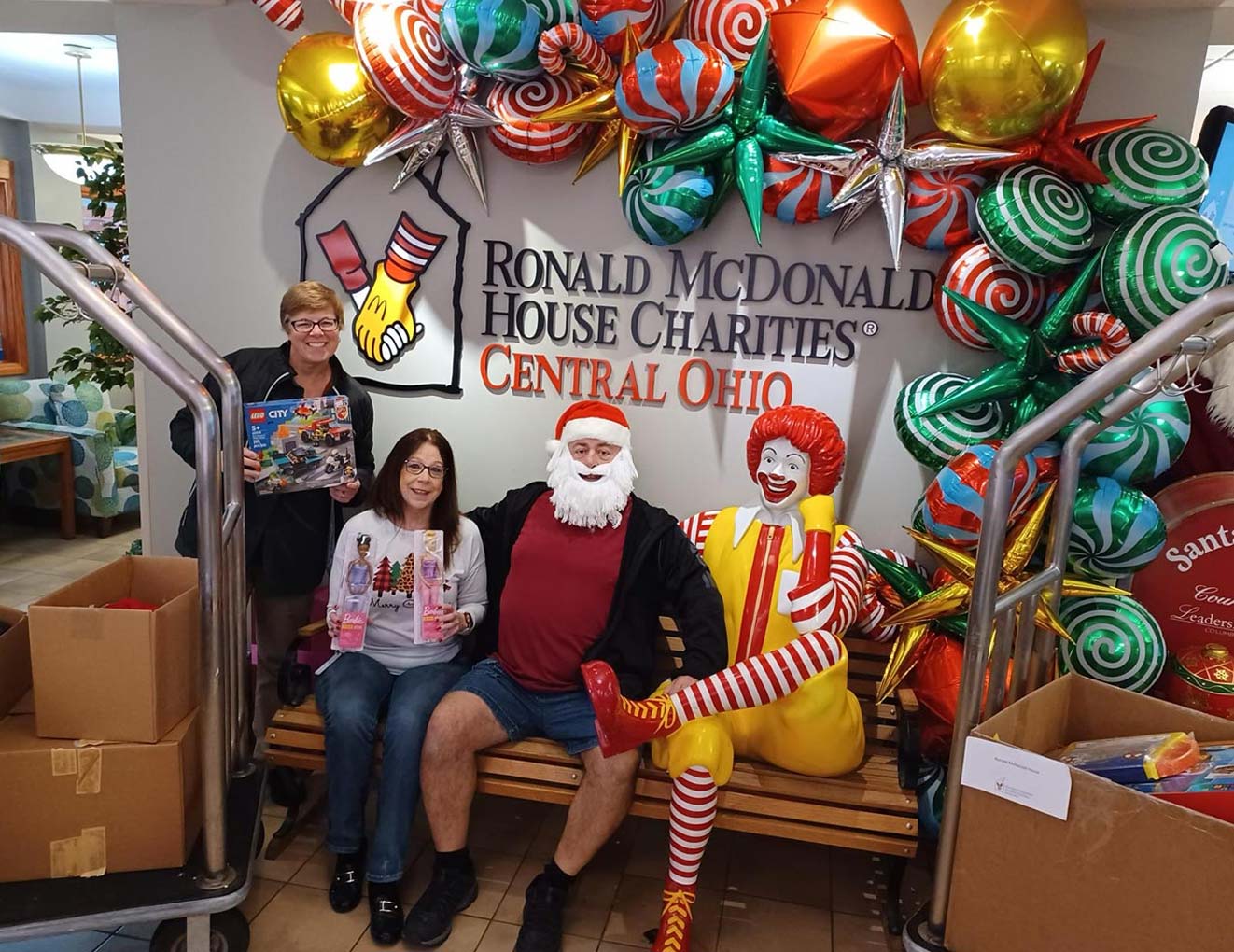 Three people pose smiling in front of a Ronald McDonald House Charities sign, surrounded by colorful holiday balloons. One person is dressed as Santa, another holds toys, and a Ronald McDonald statue sits beside them.