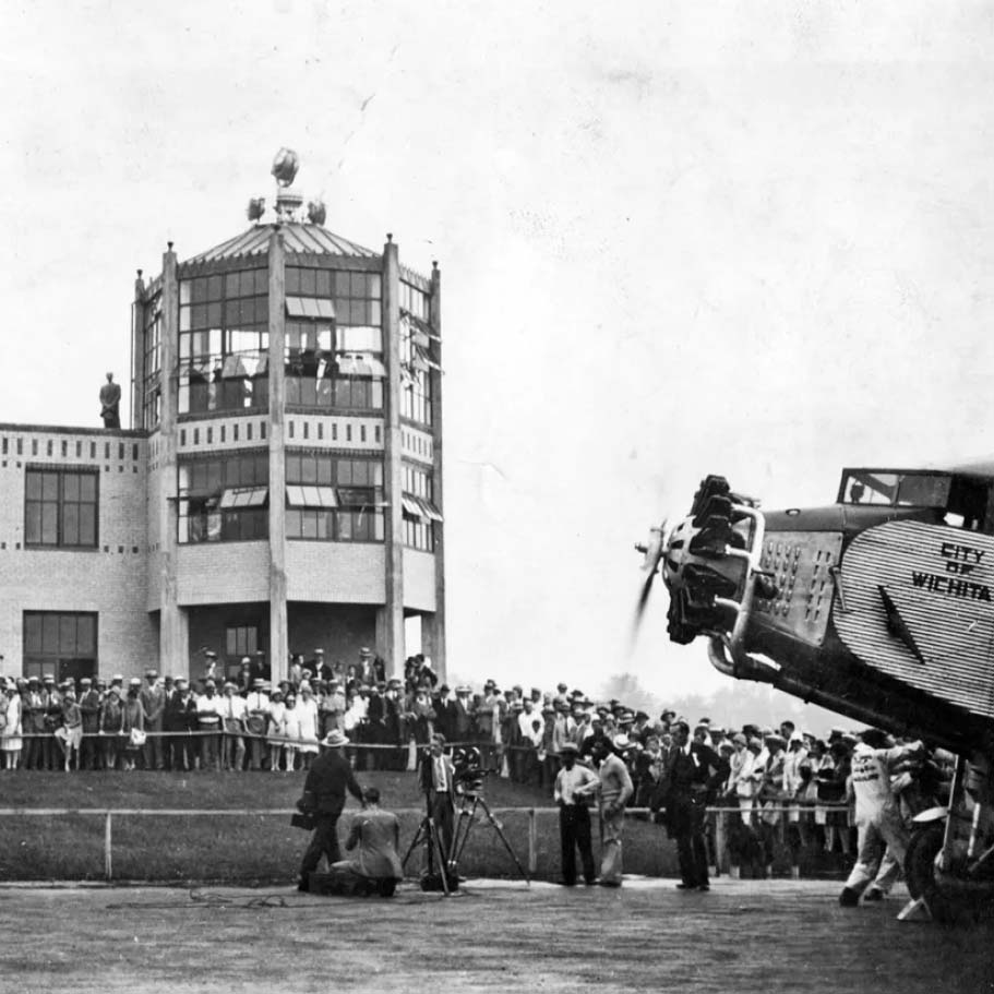A vintage airplane is parked near a crowd of people gathered outside a two-story building with large windows and a tower. Photographers and onlookers observe the scene, suggesting a significant public event.
