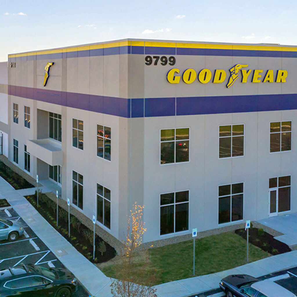 A modern Goodyear building with large windows, logo, and street number 9799 on the facade. Several parked cars are visible in the adjacent parking lot.