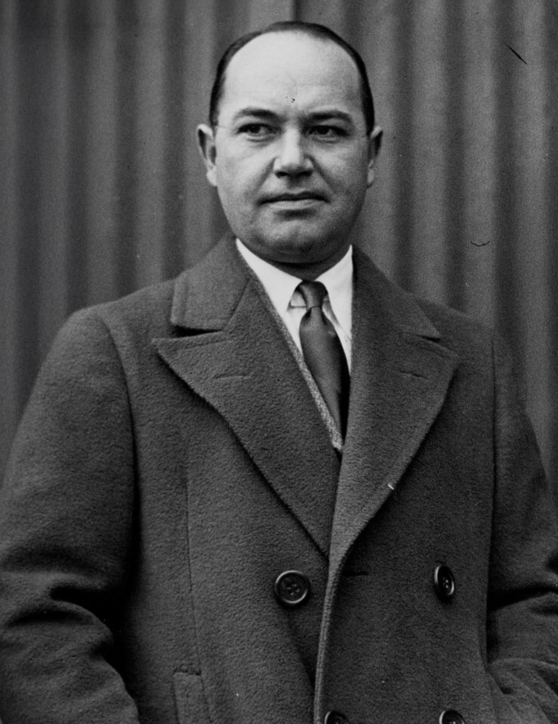 Black and white photo of a man in a double-breasted coat and tie, standing in front of a corrugated background, looking slightly to the side with a neutral expression.