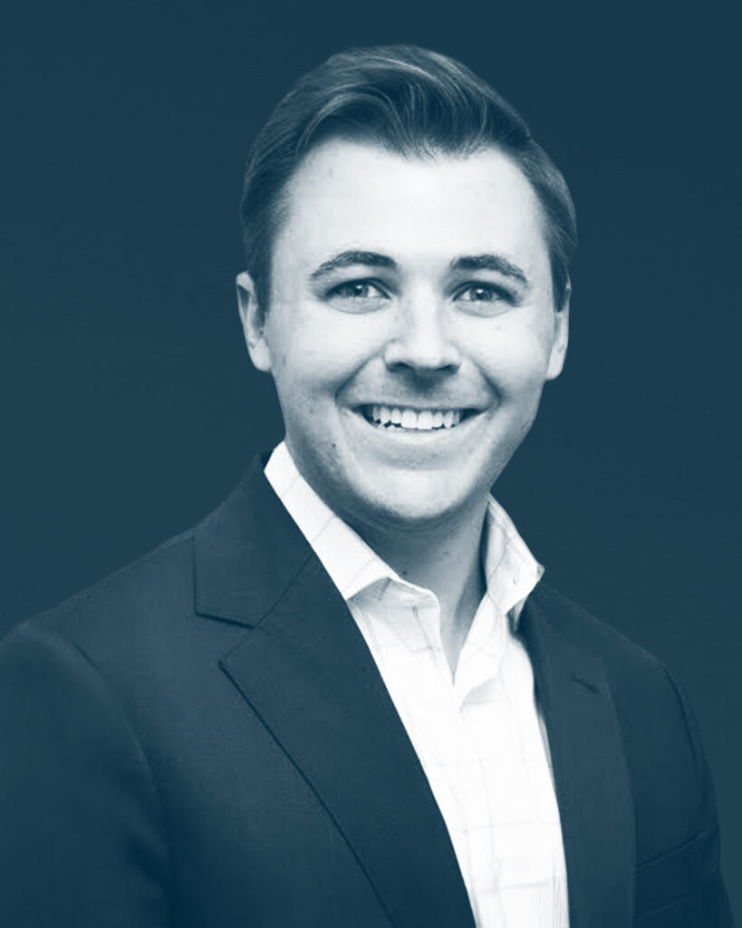 A man in a suit jacket and collared shirt smiles at the camera against a plain dark background. The image is in a blue-tinted monochrome.