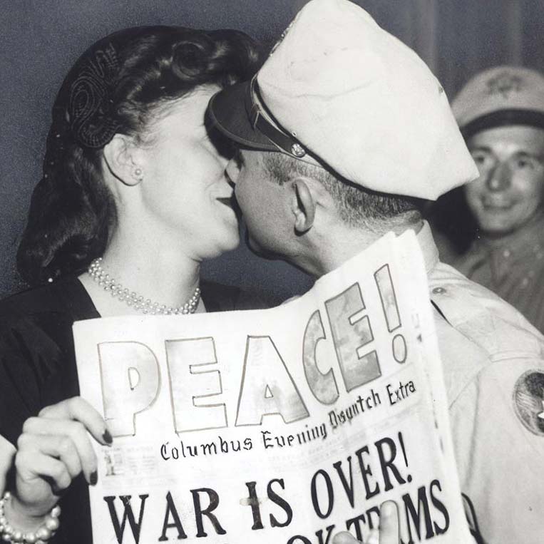 A woman and a uniformed soldier kiss while holding a newspaper with the headline PEACE! WAR IS OVER! Another smiling soldier stands in the background.
