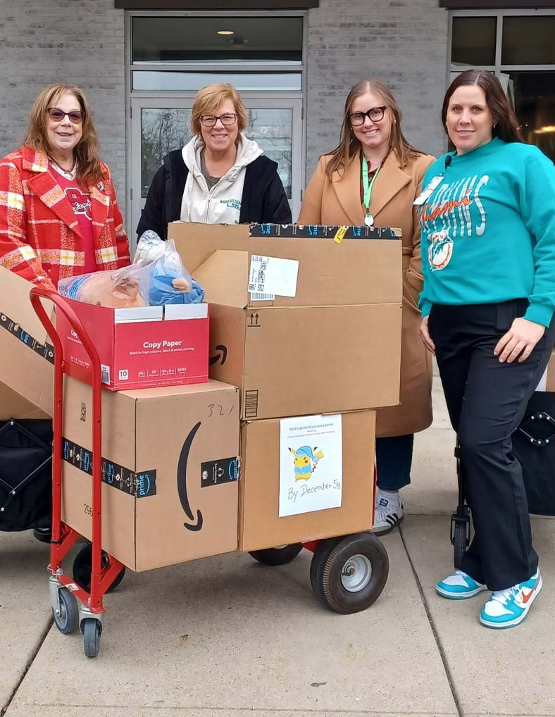 Four women stand outside a building, smiling at the camera. They are next to a red cart loaded with large cardboard boxes, bags, and other donated items. One box has a colorful drawing taped to it.