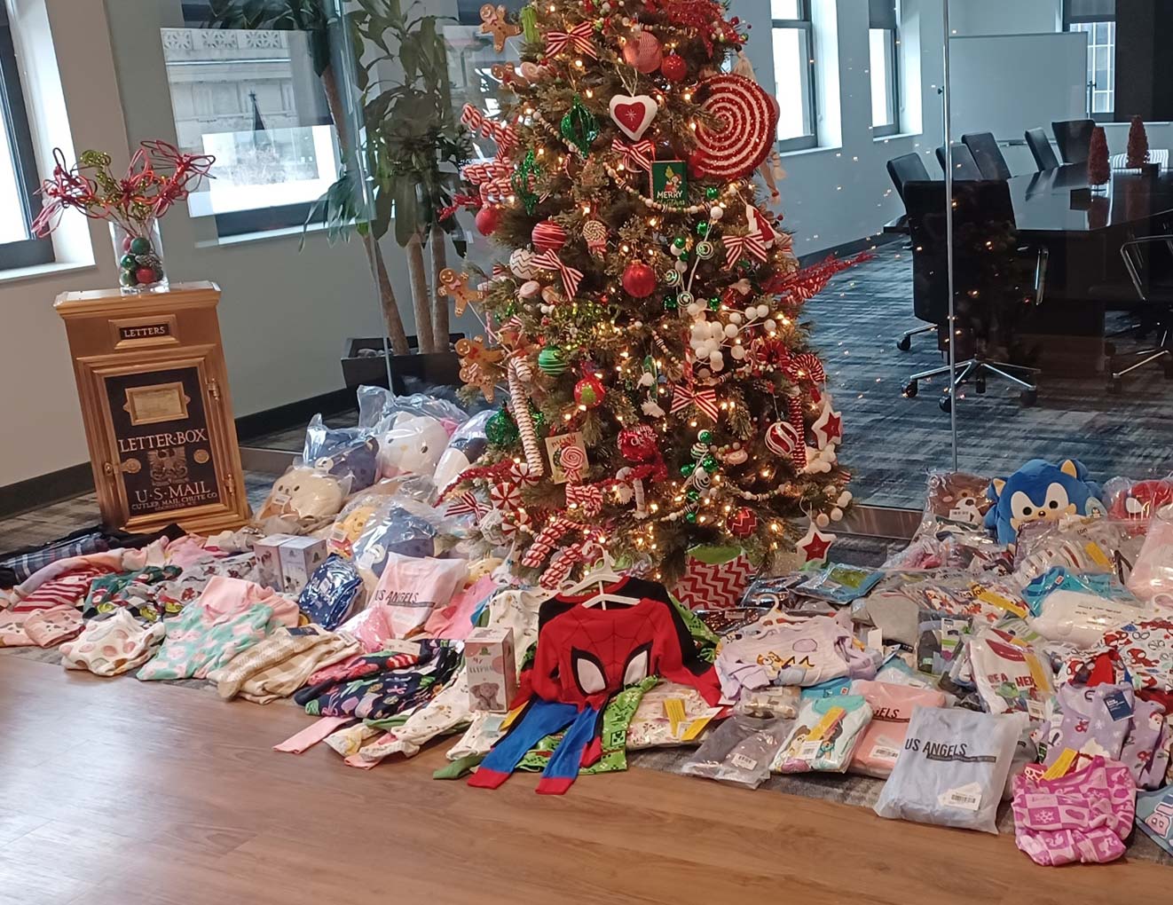 A decorated Christmas tree stands in an office with a large collection of childrens clothes, pajamas, and plush toys neatly arranged on the floor around it. A festive mailbox labeled Letters to Santa is nearby.