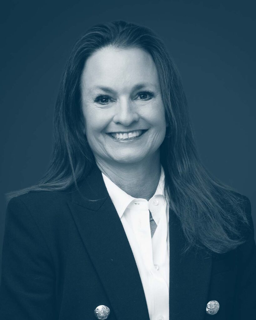 A woman with long brown hair wearing a dark blazer over a white shirt, smiling at the camera against a plain, dark background.