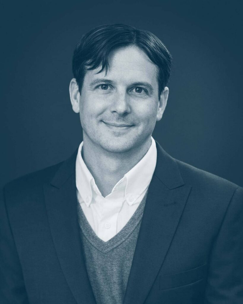 A man with short dark hair, wearing a suit jacket over a sweater and collared shirt, smiling slightly, posed against a plain dark background. The image has a blue tint.