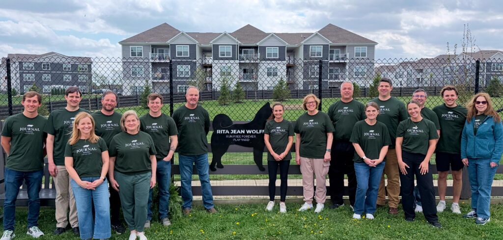 A community group wearing matching green shirts stands in front of a black dog-shaped sign reading Rita Jean Wolf Bark Park, with apartment buildings and a chain-link fence in the background.