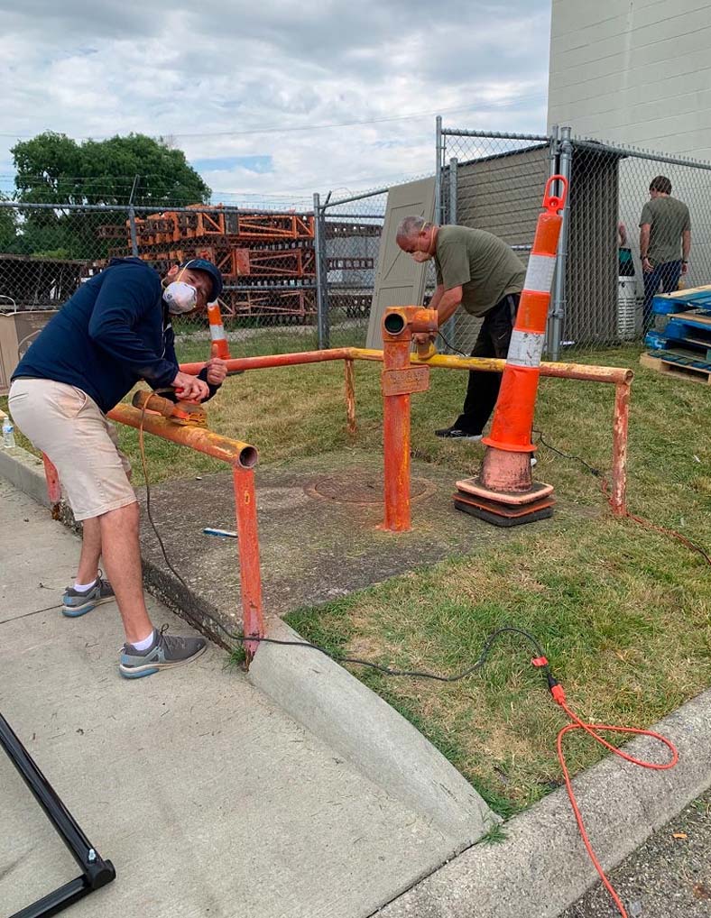 Two men in masks are sanding a rusty orange metal railing outdoors, working together as part of a community effort. One man in the foreground wears a blue long-sleeved shirt and tan shorts, while the other in green stands behind him.