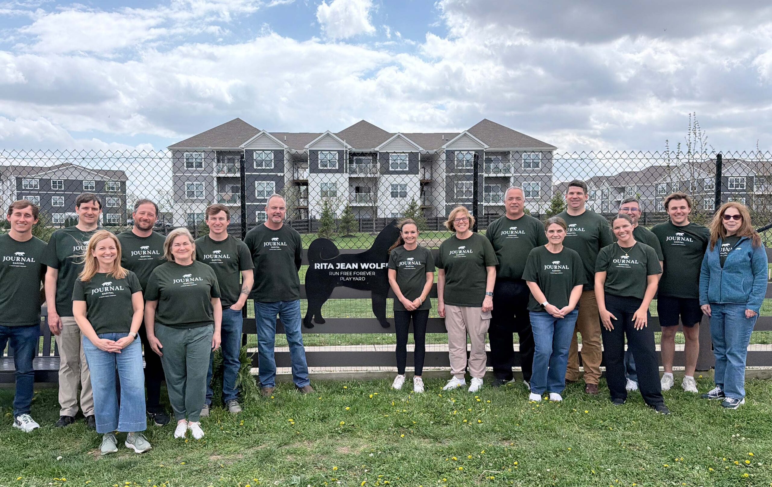 A group of people in green shirts stand in front of a sign reading Rita Jean Wolfe Memorial Park with apartment buildings and a chain-link fence in the background, under a cloudy sky.